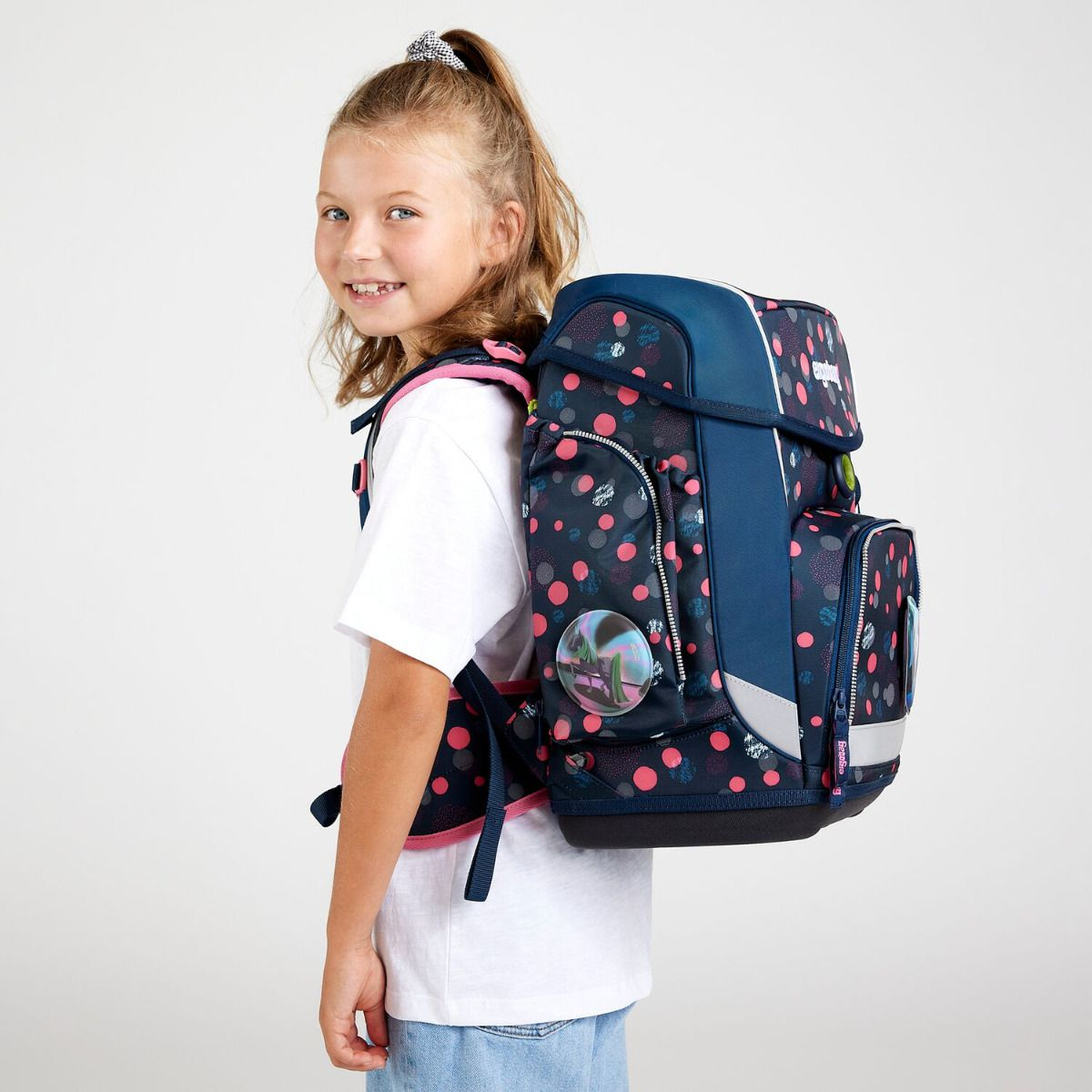 Ein Mädchen mit Pferdeschwanz lächelt und trägt ein weißes Hemd und Jeans. Sie hat einen marineblauen Rucksack mit rosa Punkten.
