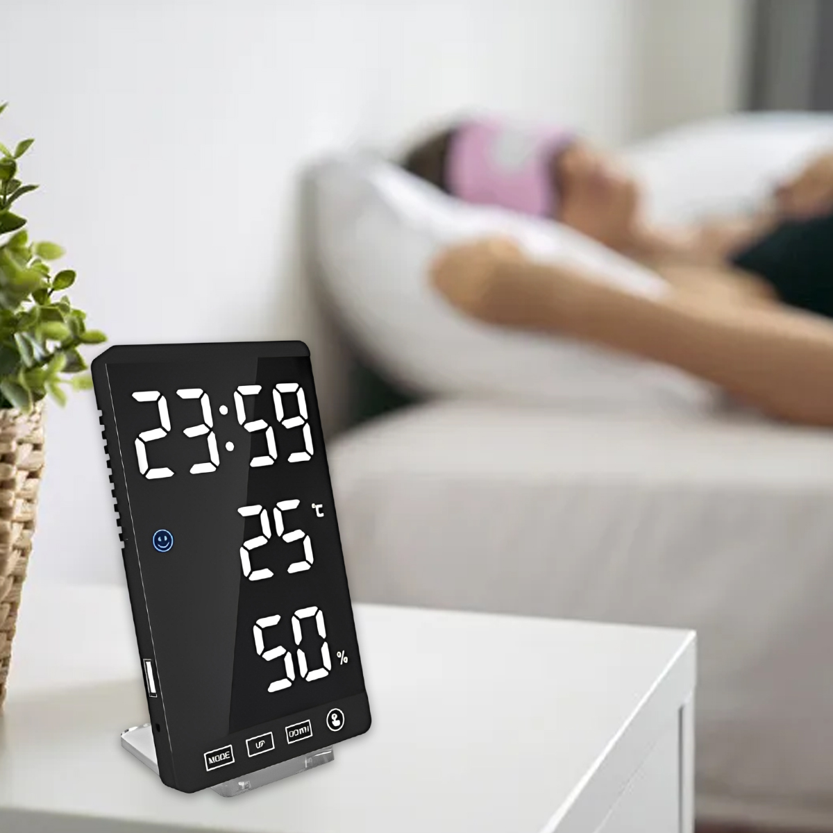 Eine Digitaluhr auf einem Nachttisch zeigt Zeit, Temperatur und Luftfeuchtigkeit an. Im Hintergrund schläft eine Person im Bett.