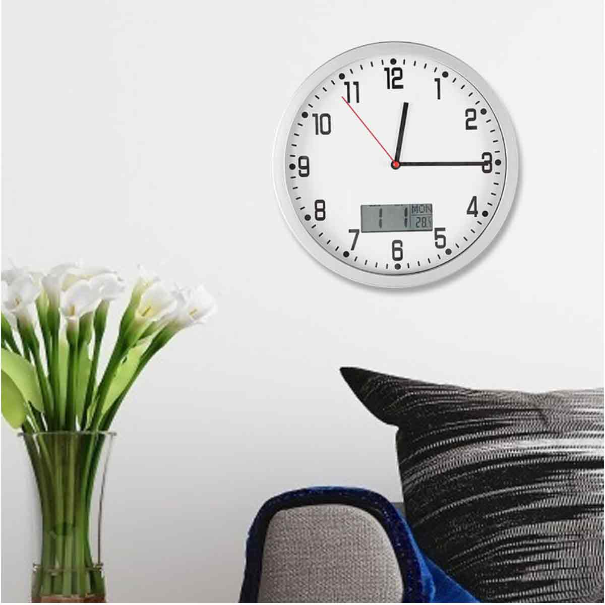 Eine weiße Uhr an einer weißen Wand. Darunter eine Vase mit weißen Blumen. Ein grauer Stuhl und ein schwarzes Kissen rechts.