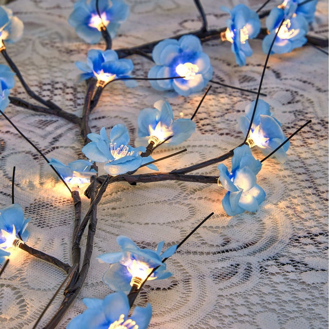 Nahaufnahme einer dekorativen Lichterkette mit blauen blütenförmigen Lichtern auf dunklen Ästen, auf weißer Spitzendecke.