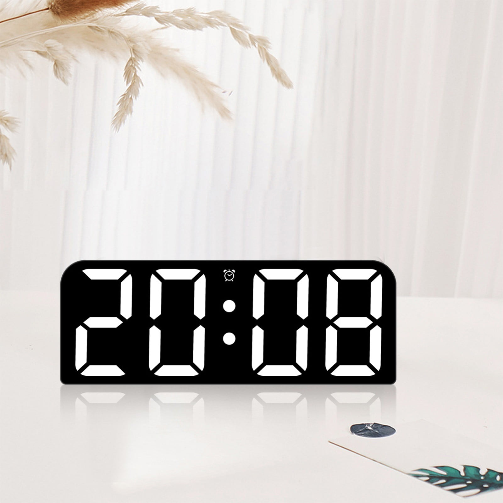 Eine Digitaluhr zeigt 20:08 auf einem schwarzen Bildschirm an. Weiße Blumen im Hintergrund, auf einem weißen Tisch.