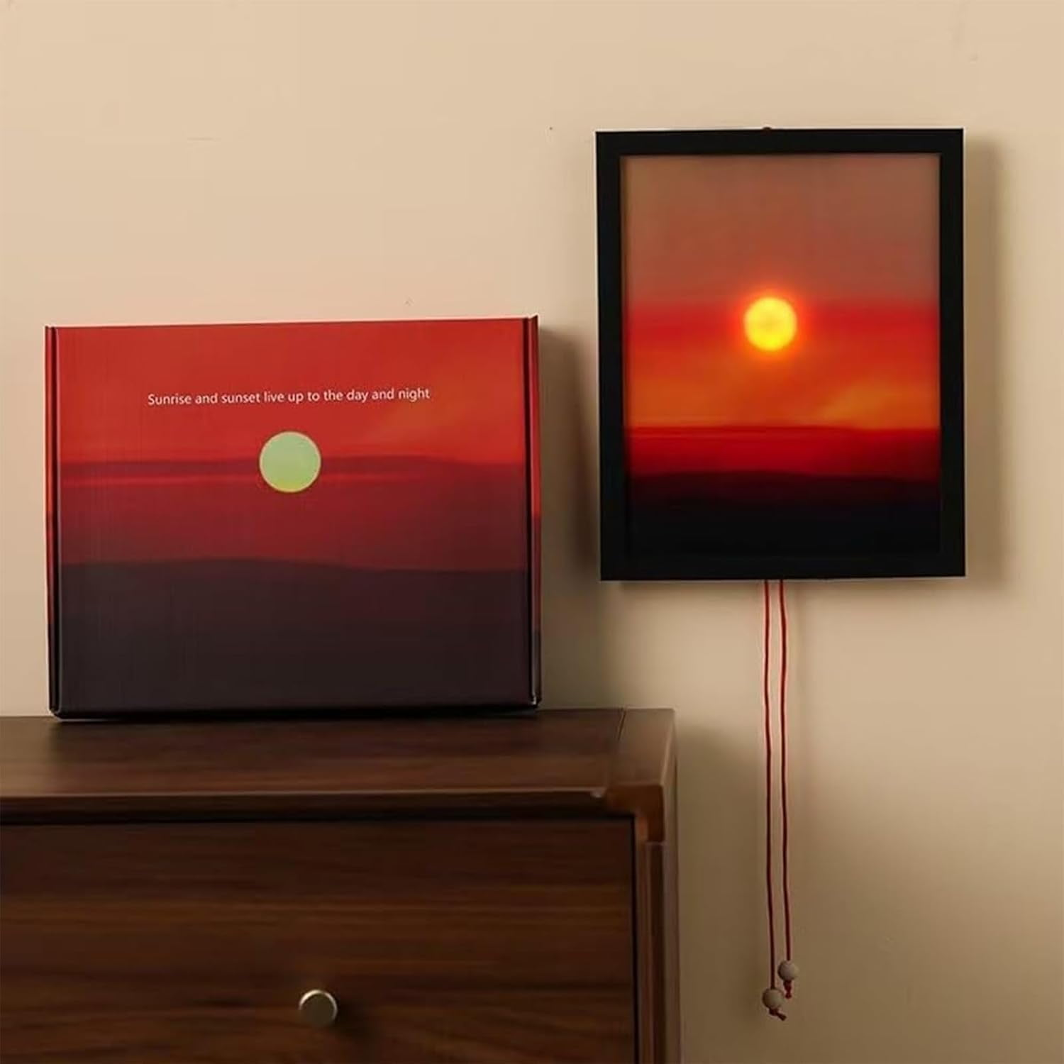 Ein gerahmtes Sonnenuntergangskunstwerk hängt neben einer Schachtel mit ähnlichem Design. Beide zeigen eine orange-rote Sonnenuntergangsszene.