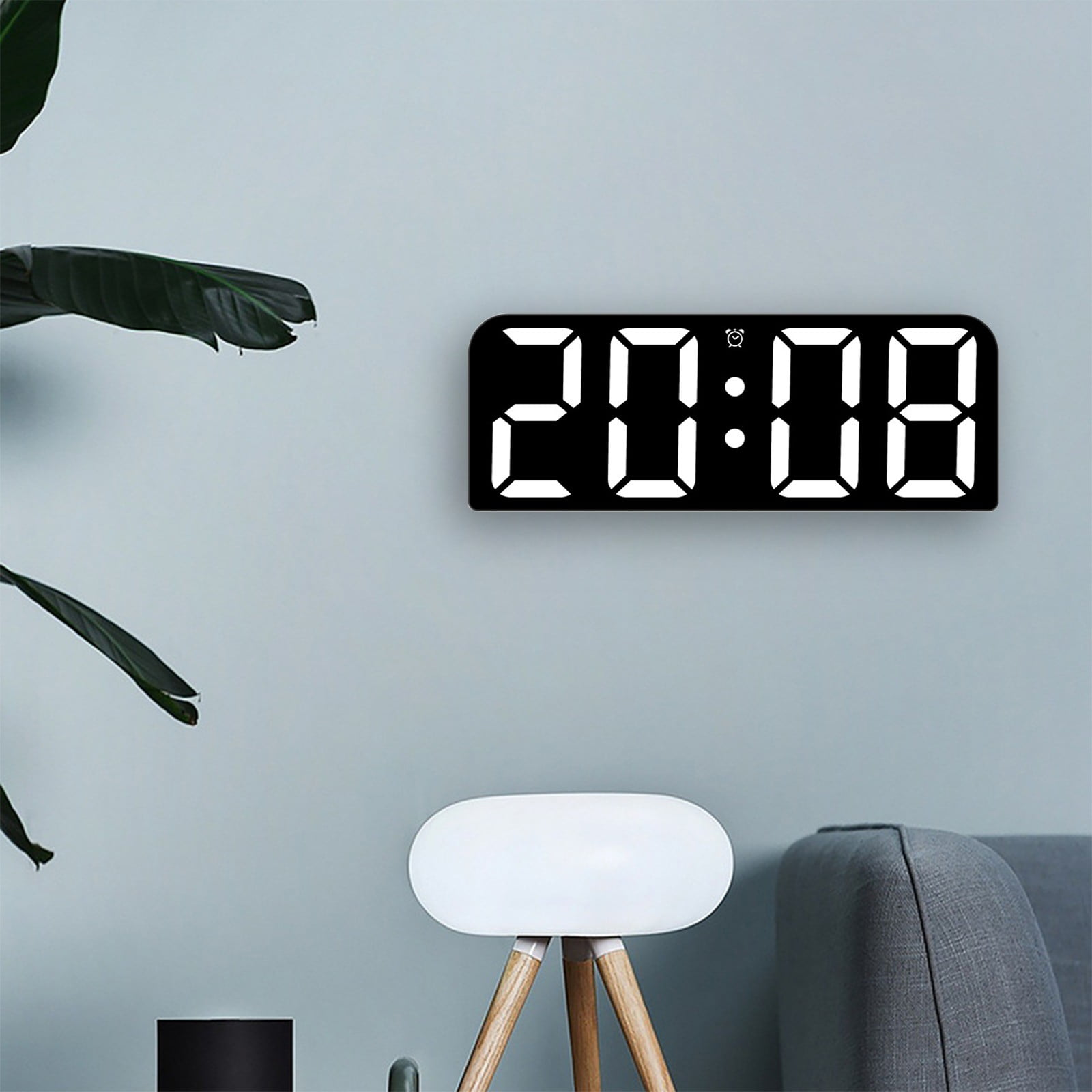Eine Digitaluhr zeigt 20:08 an der Wand an. Eine Pflanze, ein Hocker und ein Sofa befinden sich im Raum.
