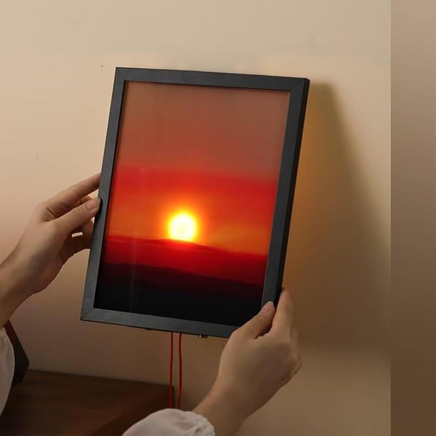 Hände halten ein gerahmtes Sonnenuntergangsbild. Die Sonne geht hinter dunklen Hügeln unter, mit roten und orangen Farben.