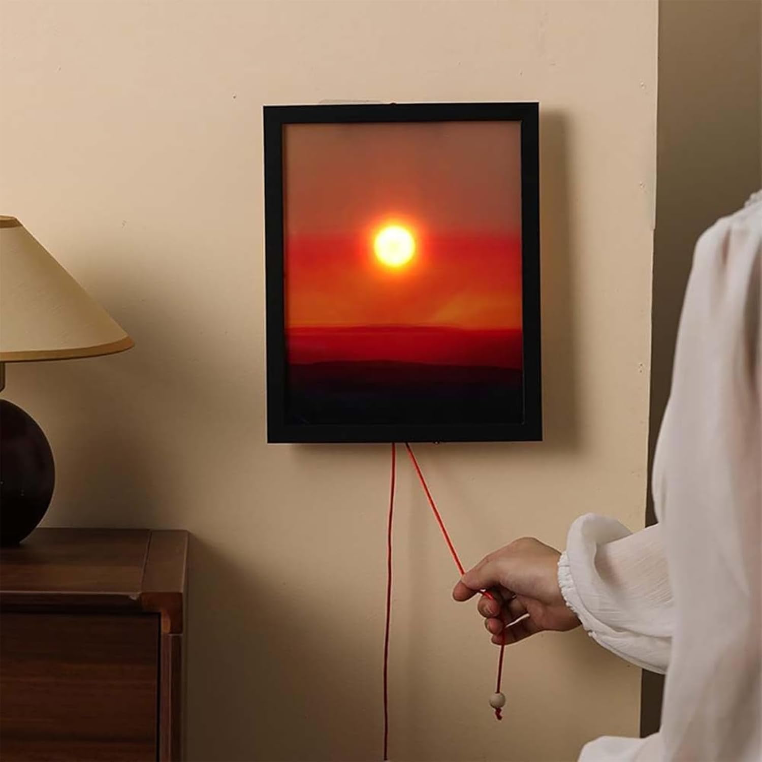 Ein gerahmtes Sonnenuntergangsbild hängt an einer Wand. Eine Person zieht an einer Schnur, die am Rahmen befestigt ist.