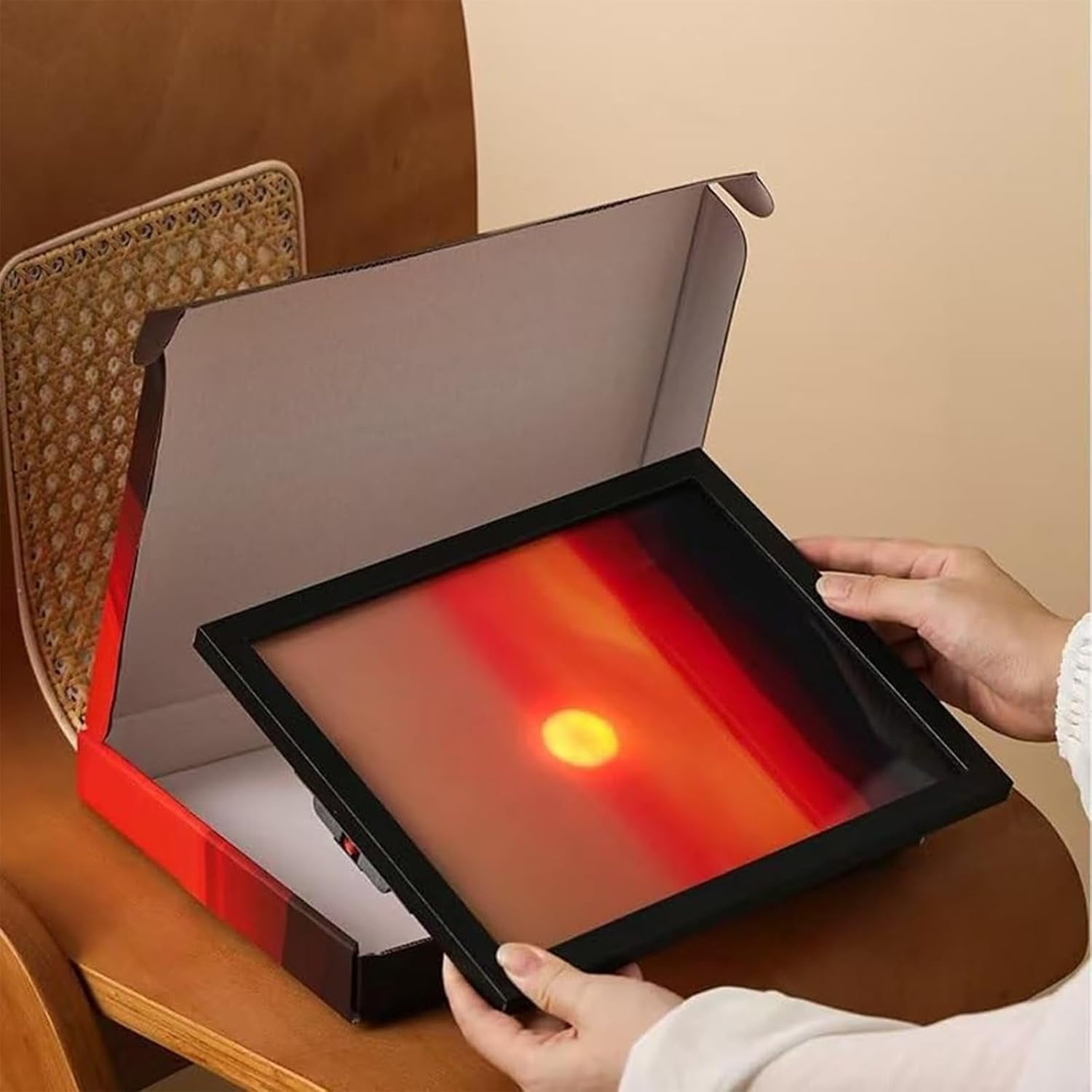 Eine Person hält ein gerahmtes Bild eines Sonnenuntergangs über einer geöffneten Box. Die Box steht auf einer Holzoberfläche.