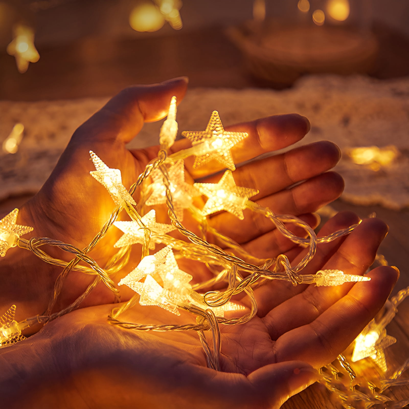 Hände halten sternförmige Lichterketten, beleuchtet. Der Hintergrund ist verschwommen mit warmen Tönen, möglicherweise Holz und Spitze.