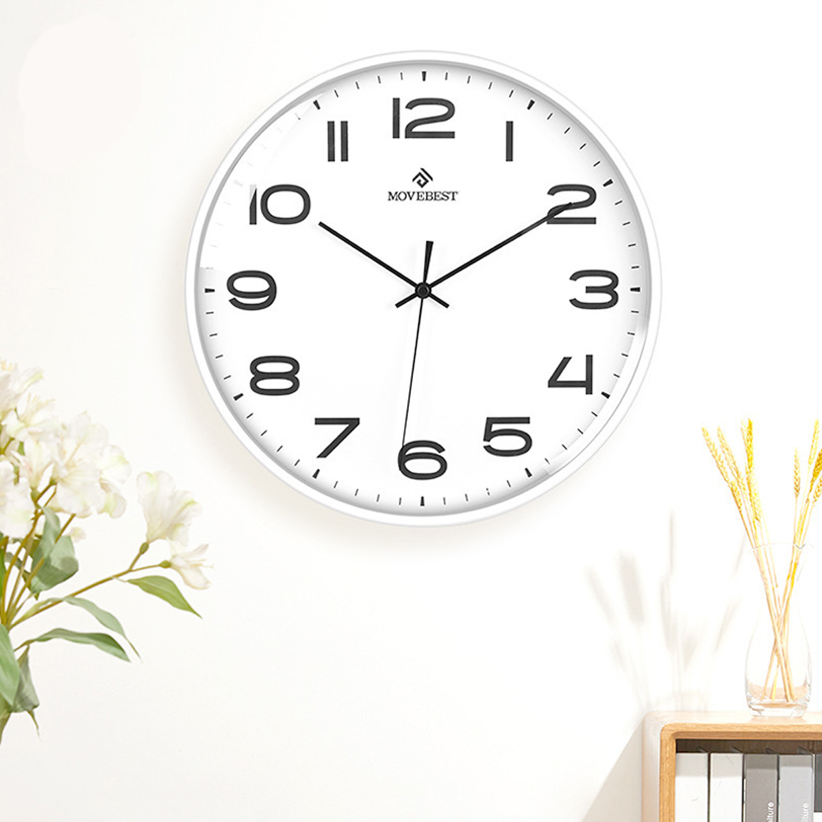 Eine weiße Uhr an einer weißen Wand. Die Uhr hat schwarze Zahlen und Zeiger. Unten sind Blumen und ein Holzregal.