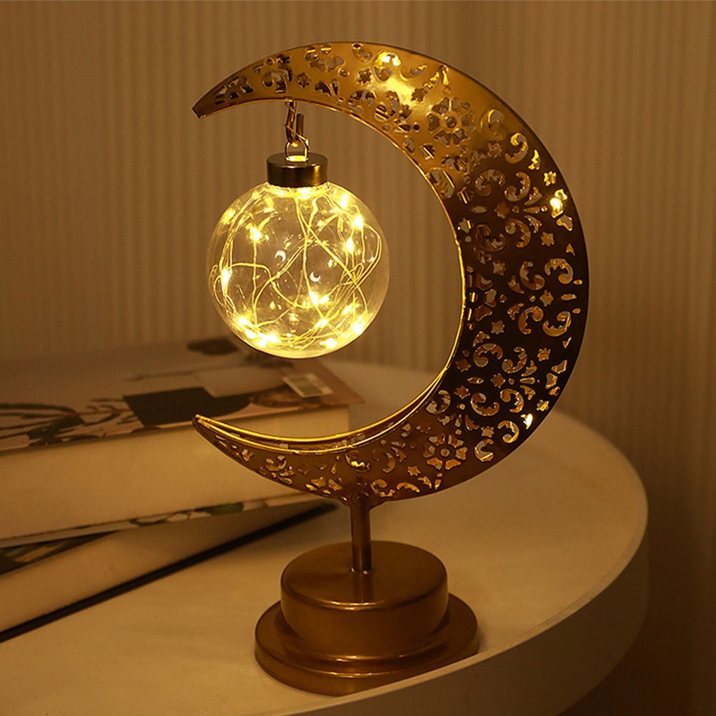 Eine goldene mondförmige Lampe mit einer Kugel mit Lichtern. Sie steht auf einem weißen Tisch mit Büchern im Hintergrund.
