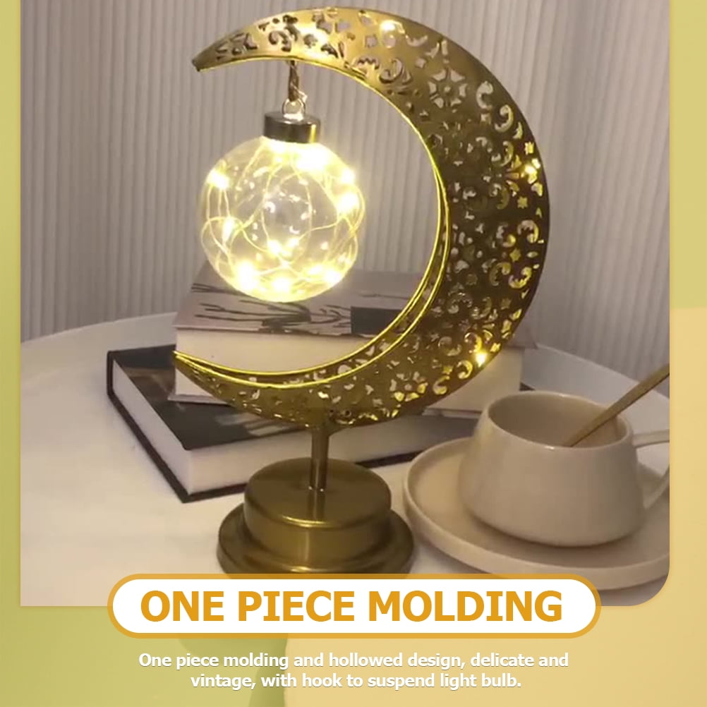 Eine goldene mondförmige Lampe mit einer leuchtenden Glühbirne. Daneben eine Tasse mit Untertasse und ein Stapel Bücher.