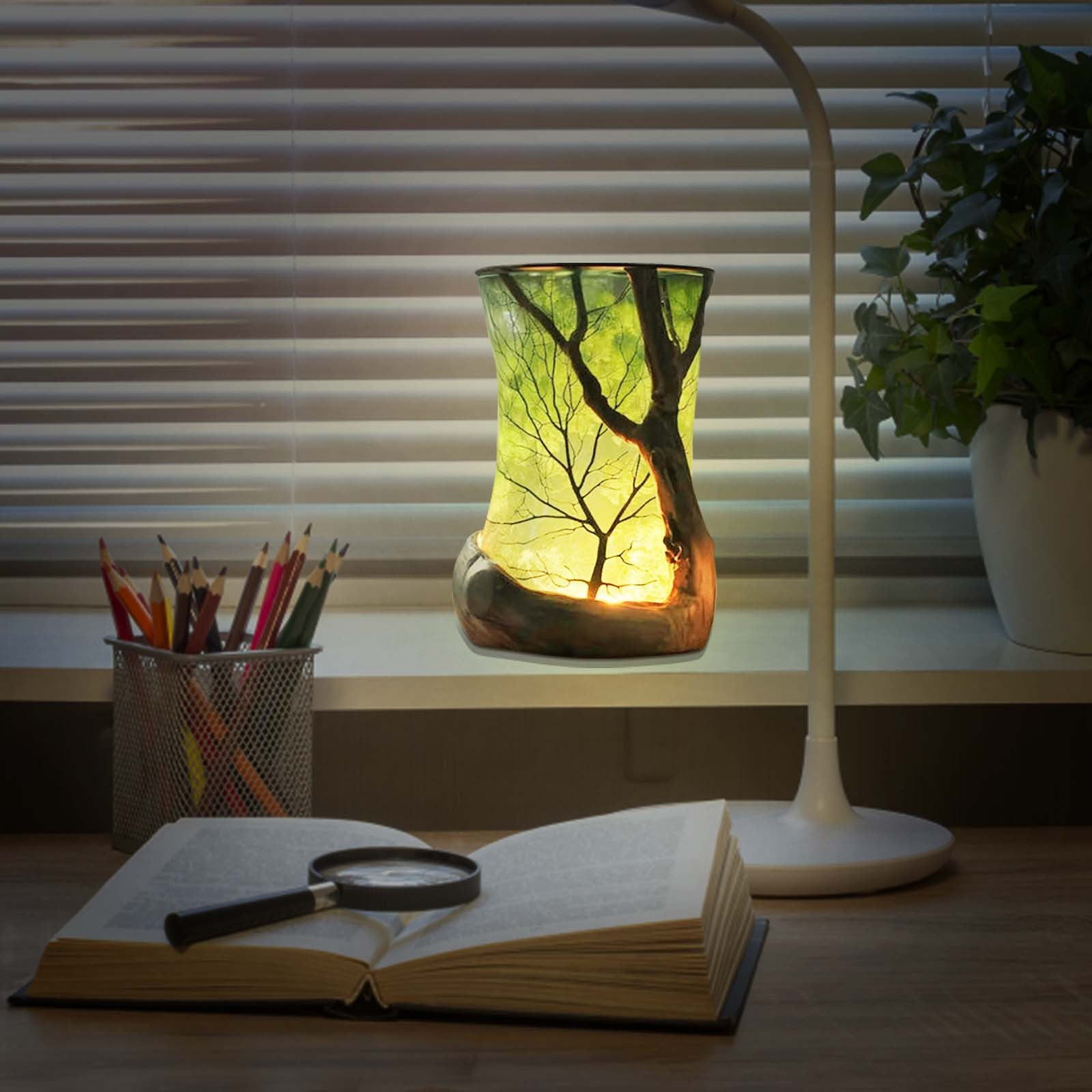 Eine beleuchtete Glaslampe mit Baumdesign, ein offenes Buch, Bleistifte und eine Schreibtischlampe auf einem Schreibtisch.