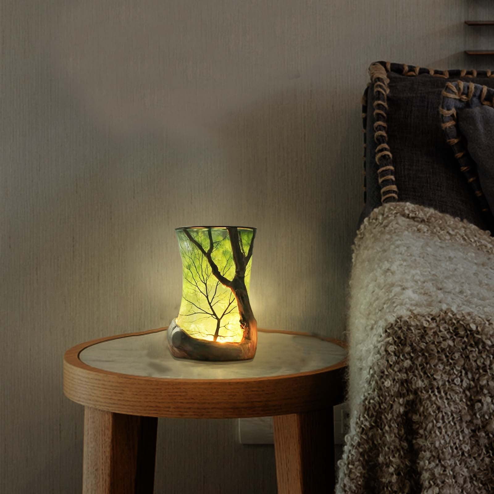 Eine beleuchtete Glaslampe mit Baumdesign steht auf einem runden Tisch. Im Hintergrund ein Bett mit einer Decke.