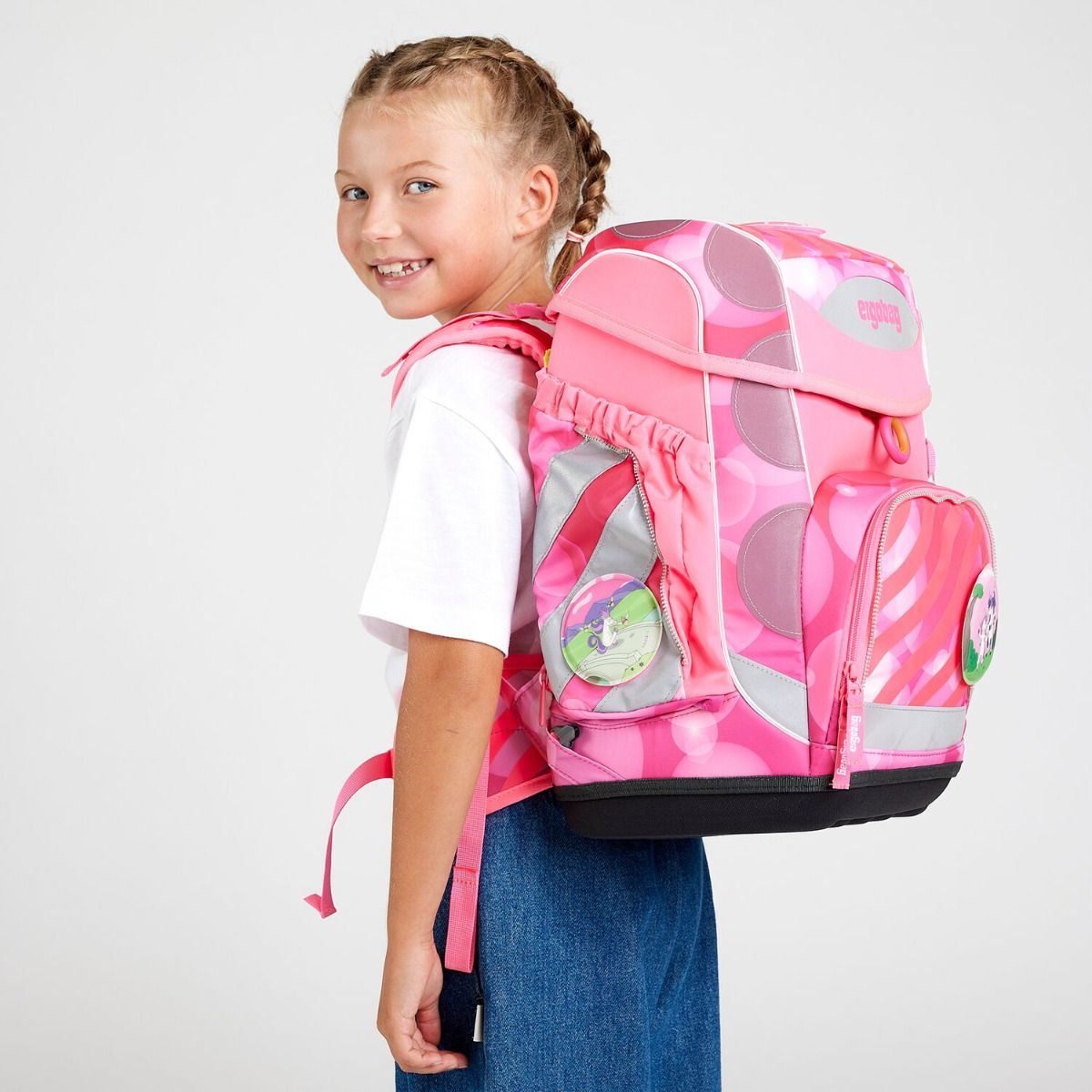 Ein lächelndes Mädchen mit Zöpfen trägt einen rosa Rucksack. Der Rucksack ist mit Kreisen und Streifen verziert. Sie trägt ein weißes Hemd und Jeans.