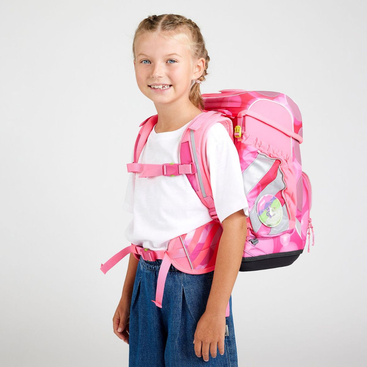 Ein lächelndes Mädchen trägt einen rosa Rucksack und Jeans. Der Rucksack hat einen reflektierenden Streifen. Sie steht vor weißem Hintergrund.