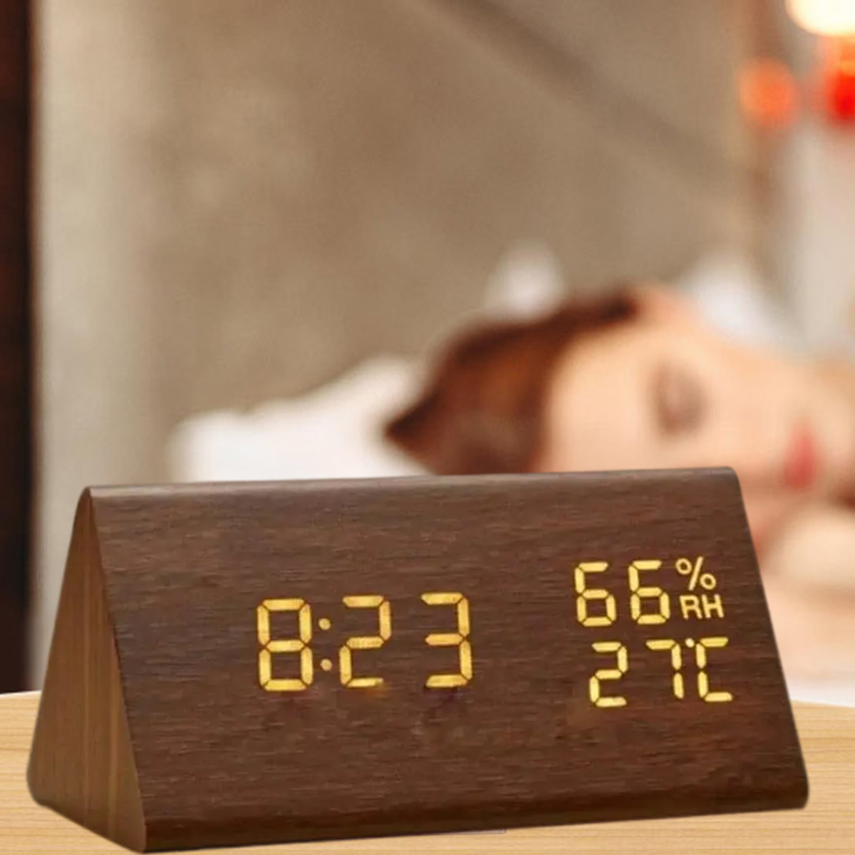 Eine hölzerne Dreiecksuhr zeigt 8:23, 66 % Luftfeuchtigkeit und 27 °C an. Im Hintergrund ist eine verschwommene Person, möglicherweise schlafend.