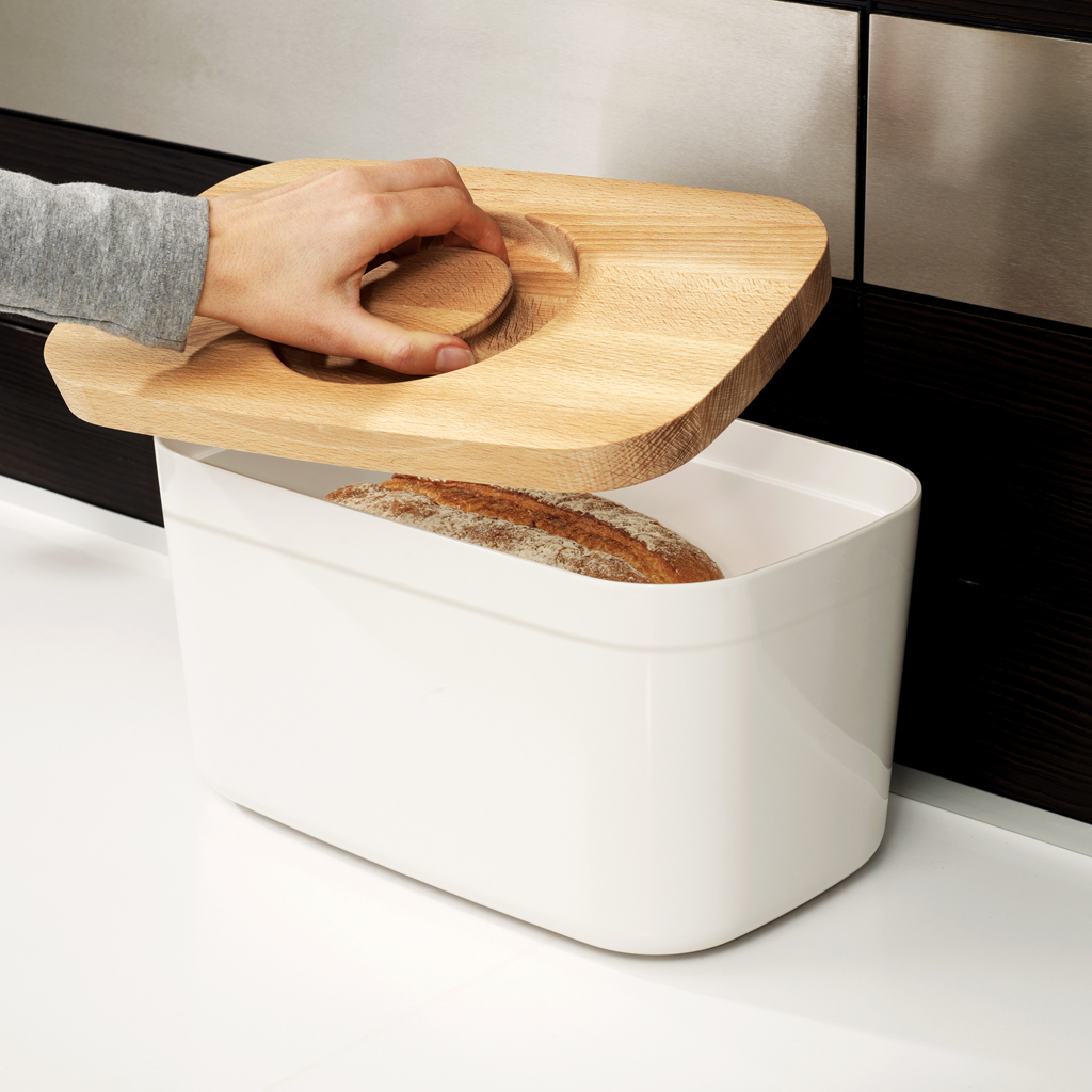Eine Hand hebt einen hölzernen Brotdeckel. Im Inneren liegt ein Brotlaib. Die Box ist weiß, auf einer weißen Oberfläche.