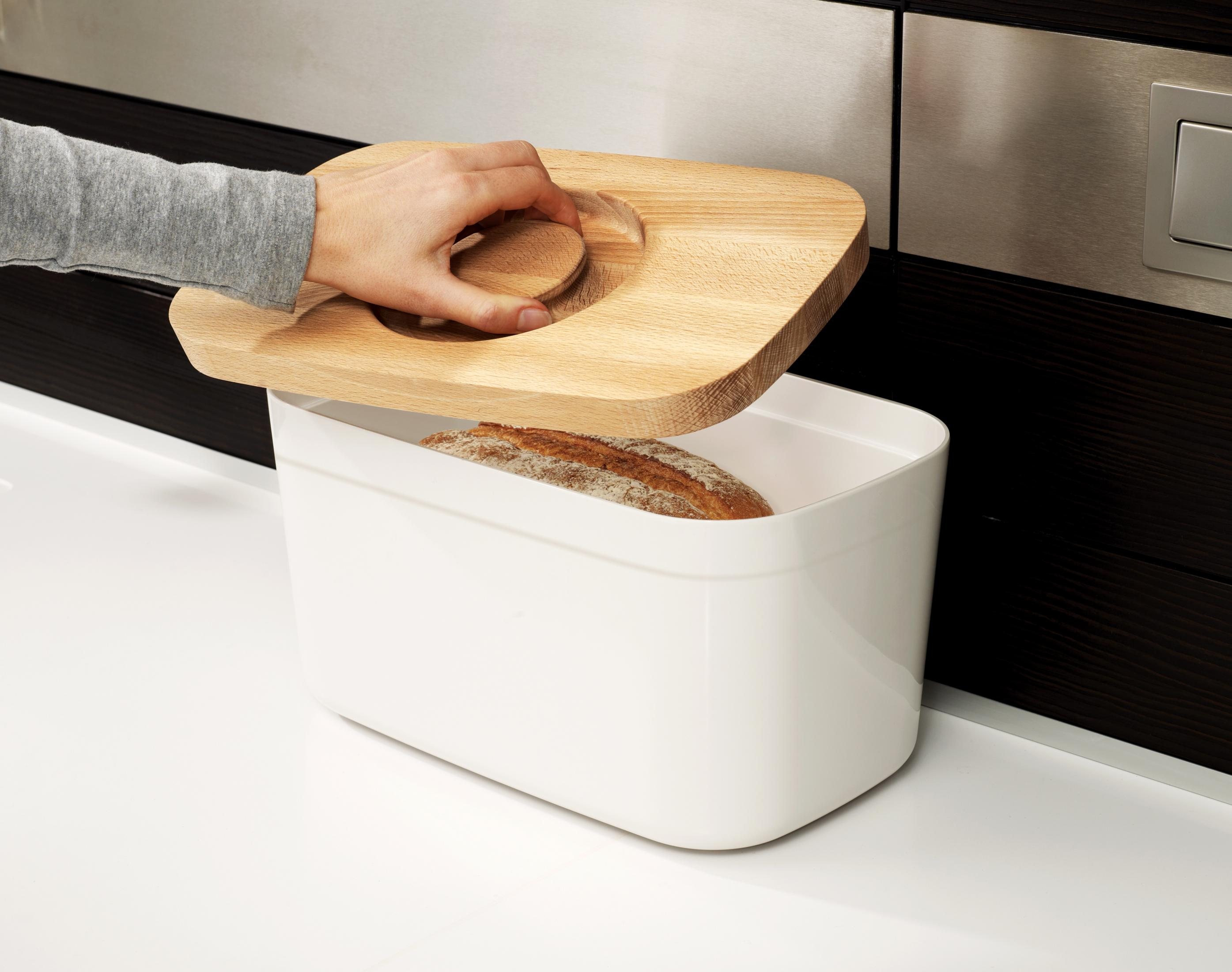 Eine Hand hebt einen Holzdeckel einer Brotdose. Darin befindet sich ein Brotlaib. Die Box ist weiß.