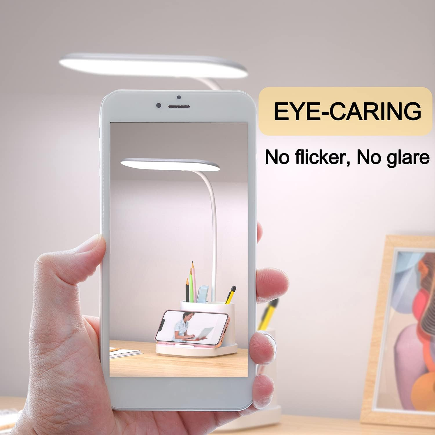 Eine Hand hält ein Telefon, das eine Schreibtischlampe zeigt. Die Lampe ist weiß, mit Bleistiften, einem Telefon und einem Bilderrahmen.