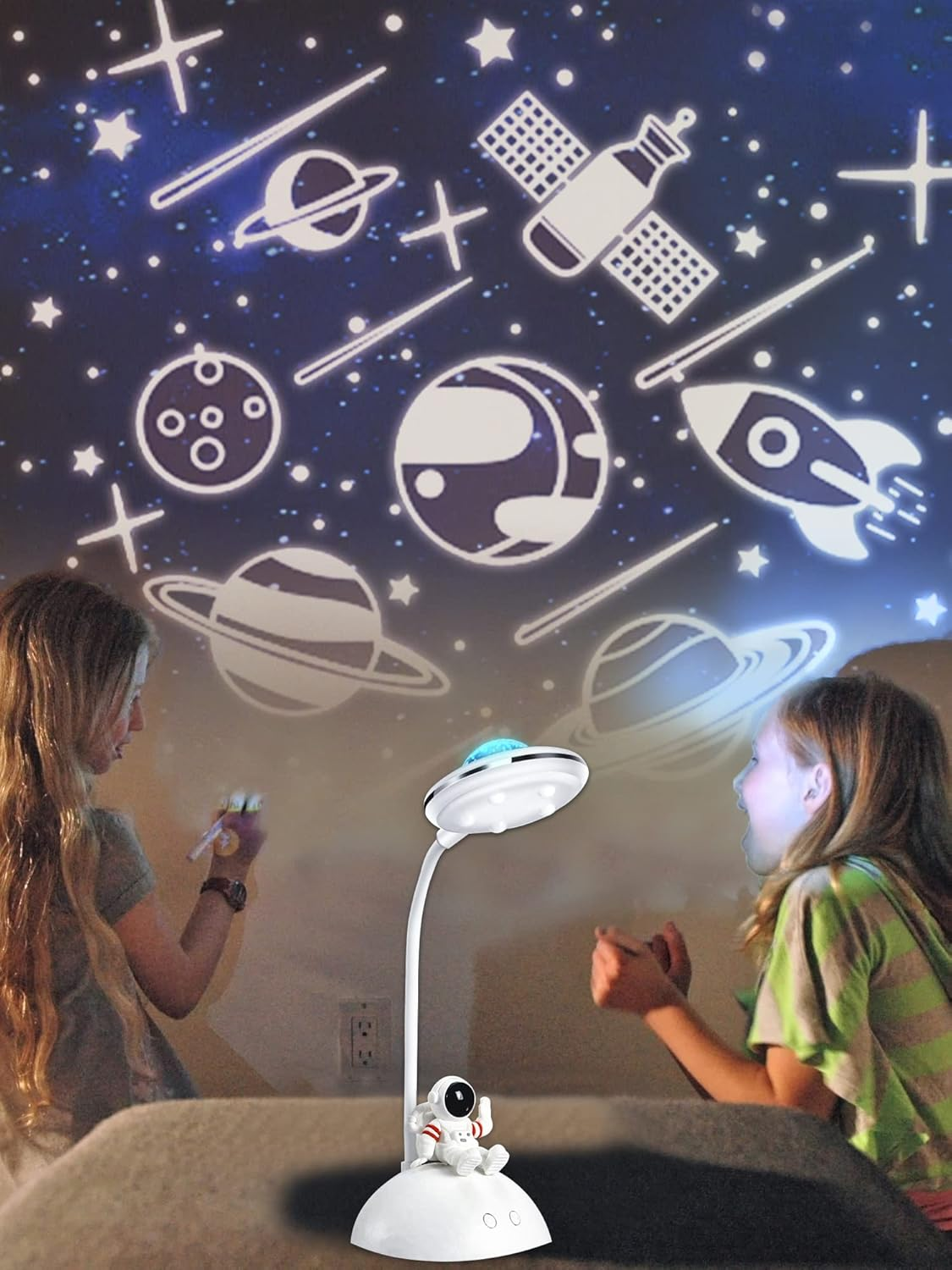 Zwei Mädchen betrachten einen Sternenhimmel, der von einer Lampe projiziert wird. Die Lampe zeigt eine Rakete, Satellit, Planeten und eine Astronautenfigur.