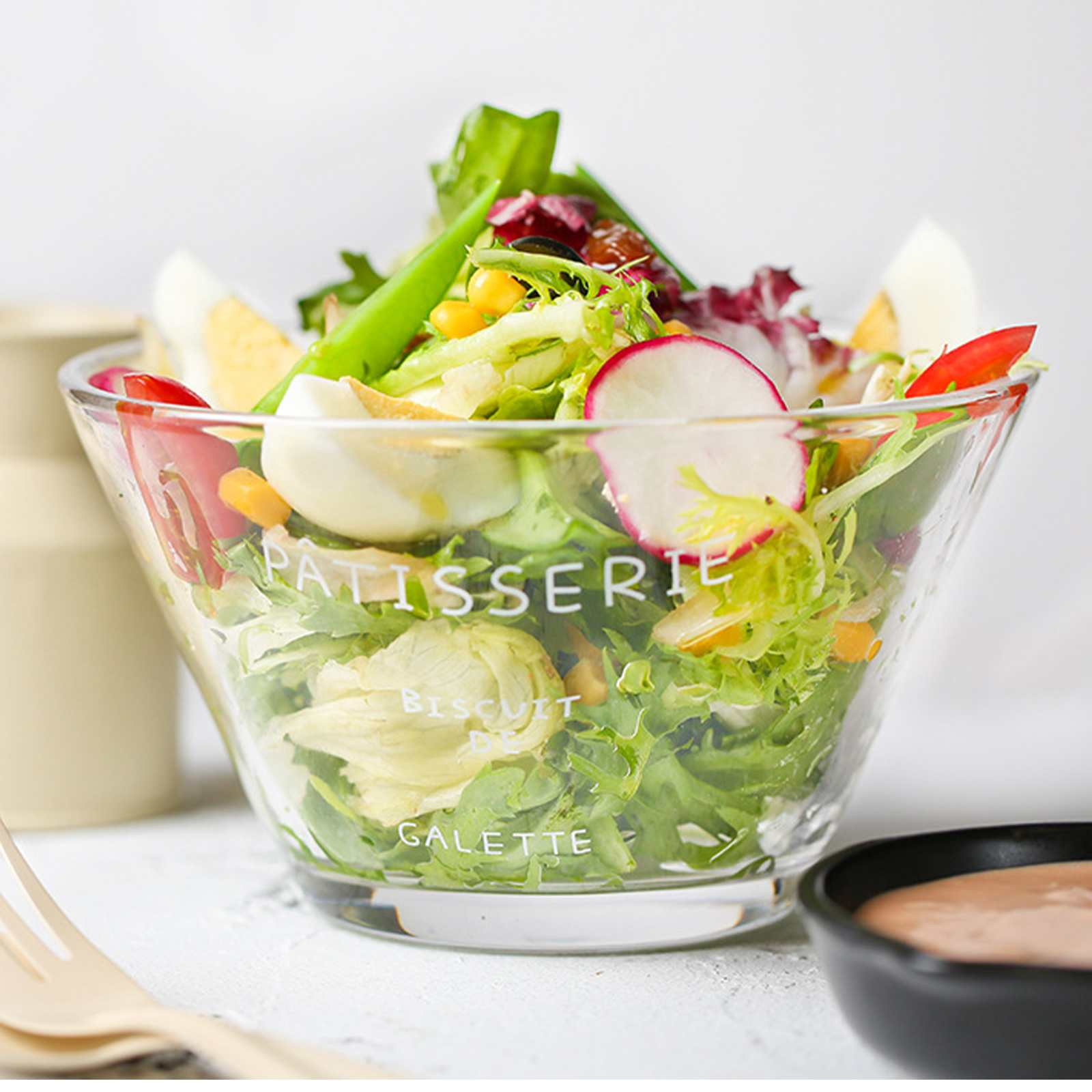 Eine Glasschüssel mit Salat, darunter Salat, Radieschen, Eier und Tomaten. Rechts eine schwarze Schüssel mit Soße.
