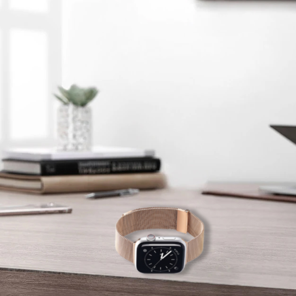 Eine Apple Watch mit Mesh-Armband liegt auf einem Holztisch. Bücher, ein Stift und eine Pflanze befinden sich im Hintergrund.