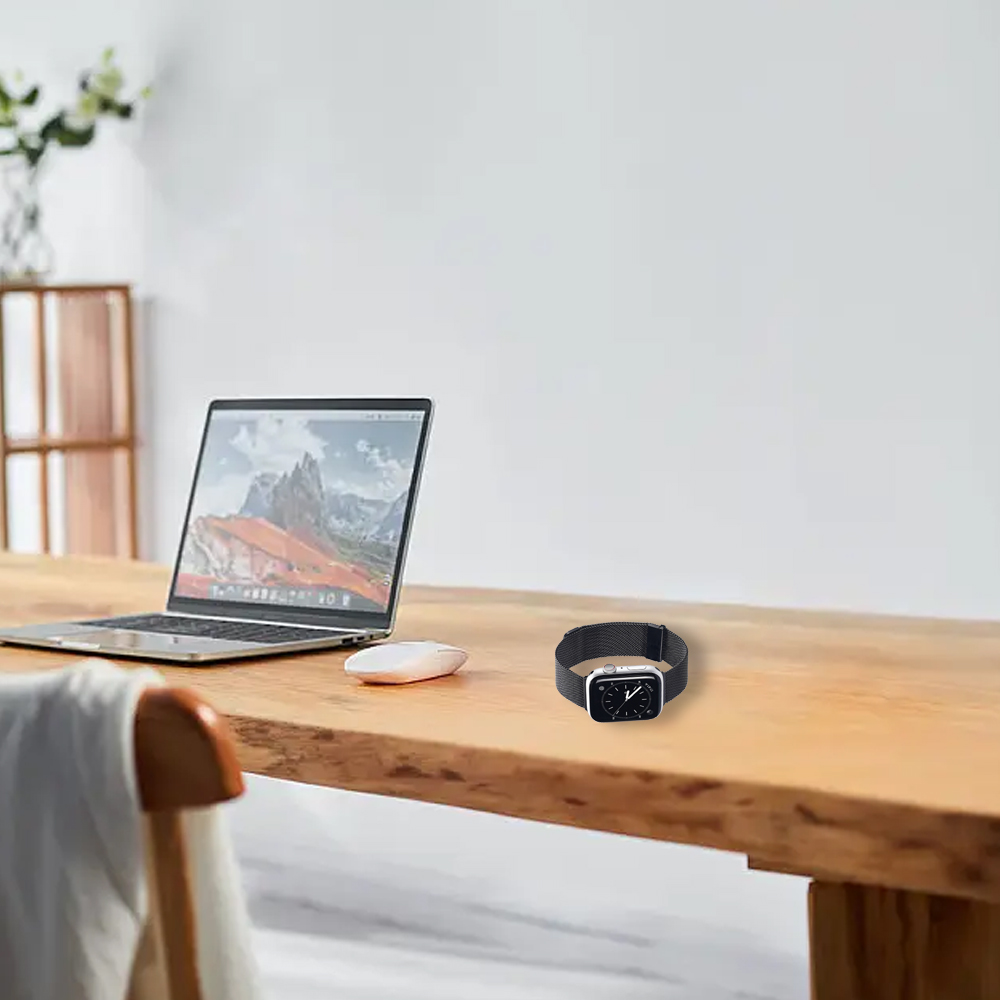 Auf einem Holztisch liegen ein Laptop, eine Maus und eine Smartwatch. Im Vordergrund steht ein Stuhl mit weißem Tuch. Weiße Wände im Hintergrund.