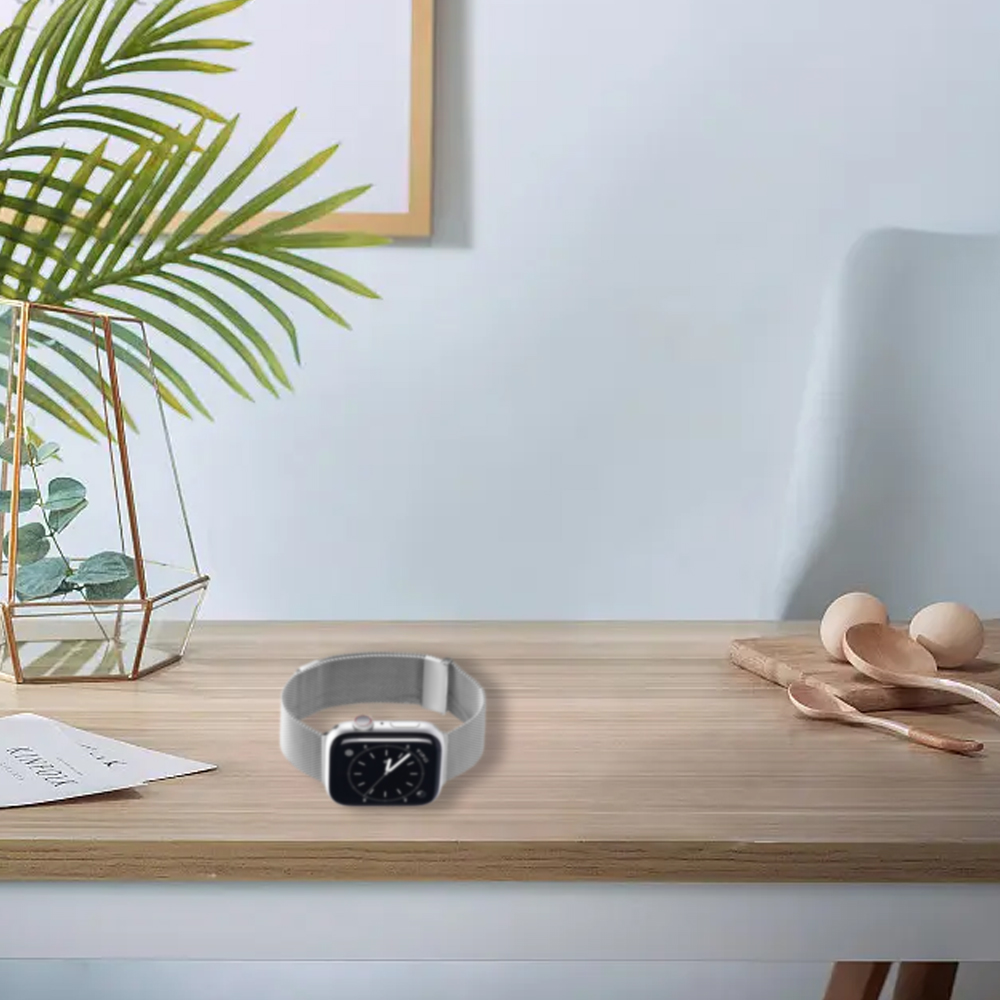 Eine silberne Apple Watch liegt auf einem Holztisch. Ein Glasterrarium, grüne Blätter und Holzlöffel befinden sich ebenfalls auf dem Tisch.