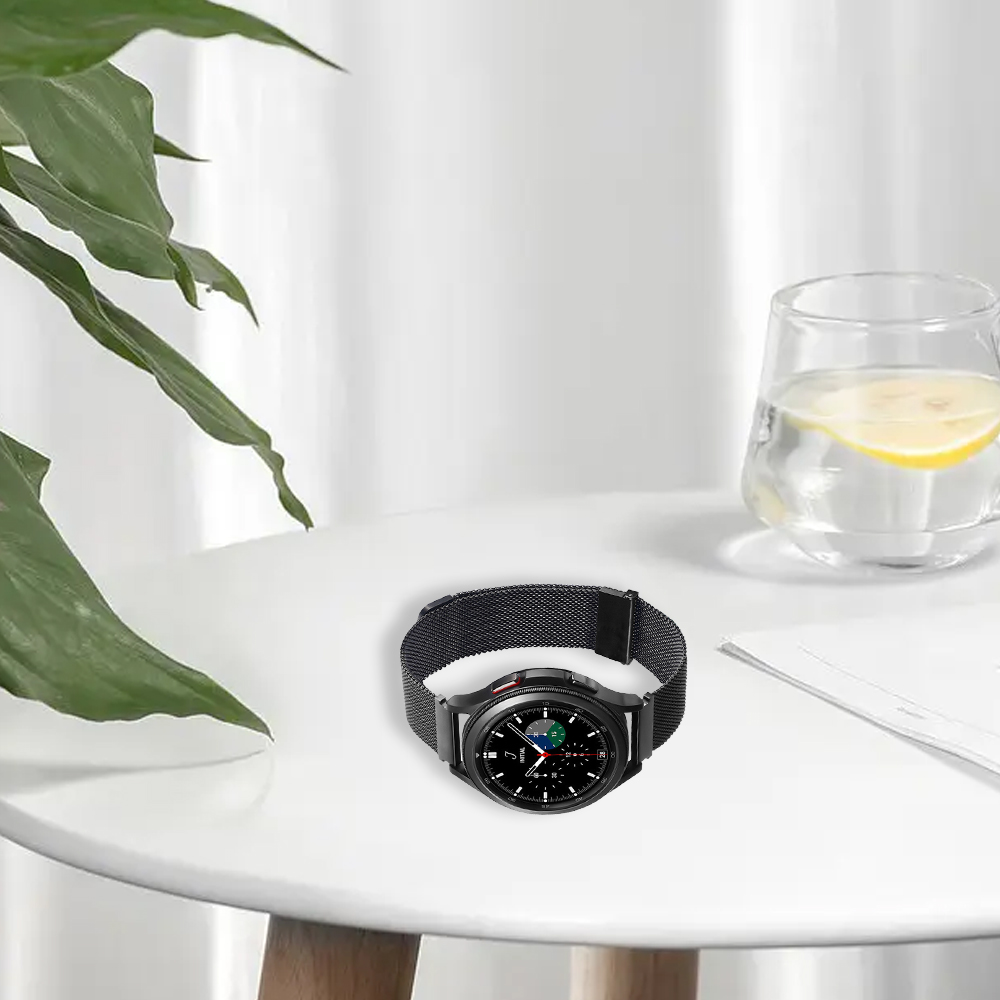 Eine schwarze Smartwatch mit Mesh-Armband liegt auf einem weißen Tisch. Ein Glas Wasser mit Zitrone und eine Pflanze sind daneben.