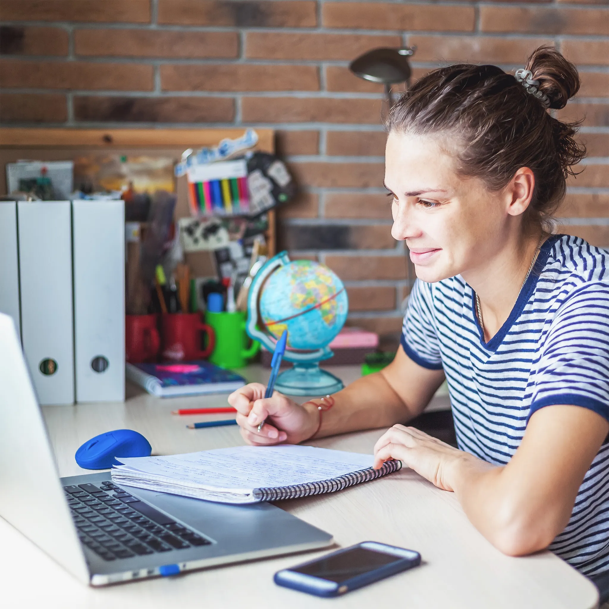 Frau im gestreiften Hemd schreibt in ein Notizbuch. Laptop, Telefon und Globus stehen auf dem Schreibtisch. Ziegelmauer im Hintergrund.