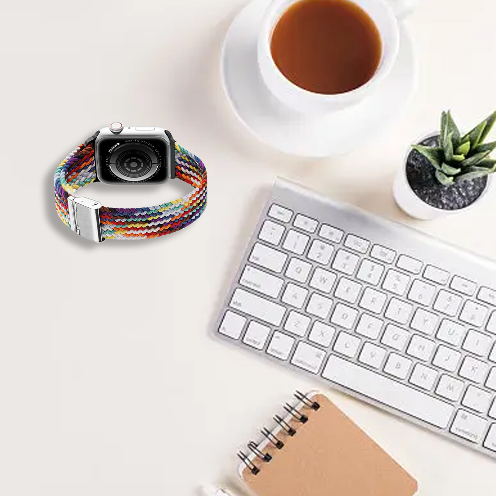 Eine Apple Watch mit buntem geflochtenem Armband, Tastatur, Kaffeetasse und kleiner Pflanze auf weißer Oberfläche.