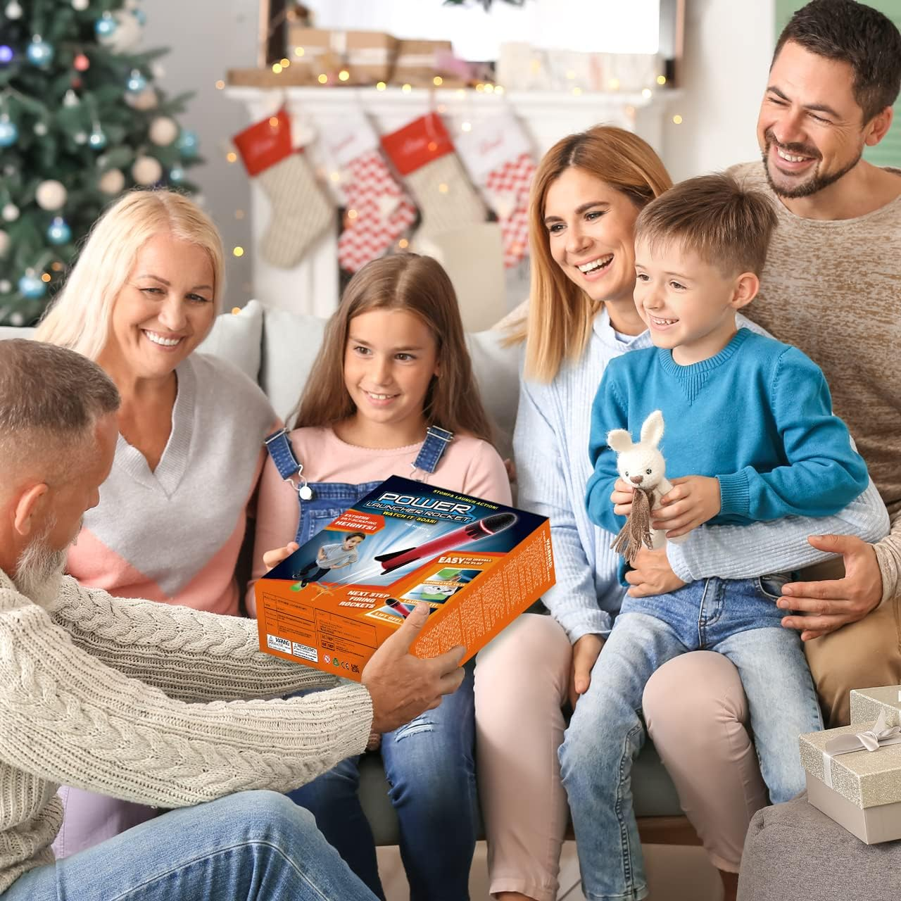 Eine Familie versammelt sich, um ein Geschenk zu öffnen. Ein Mann präsentiert der lächelnden Familie eine Raketenwerfer-Spielzeugkiste. Weihnachtsbaum und Kamin im Hintergrund.