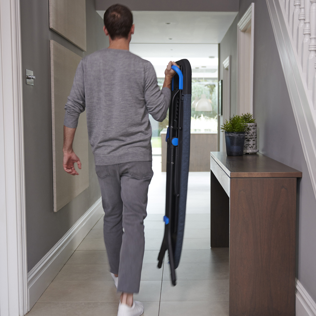 Un homme marche dans un couloir portant une planche à repasser pliée. Il porte des vêtements gris. Il y a une table en bois et des plantes à droite.
