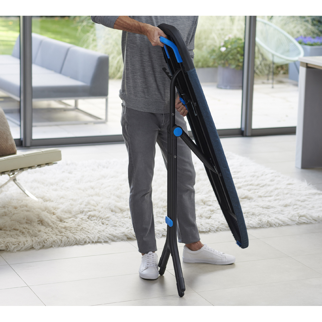 Une personne tient un banc pliant noir et bleu. L'arrière-plan montre un tapis blanc, un canapé et un jardin visible par une porte vitrée.