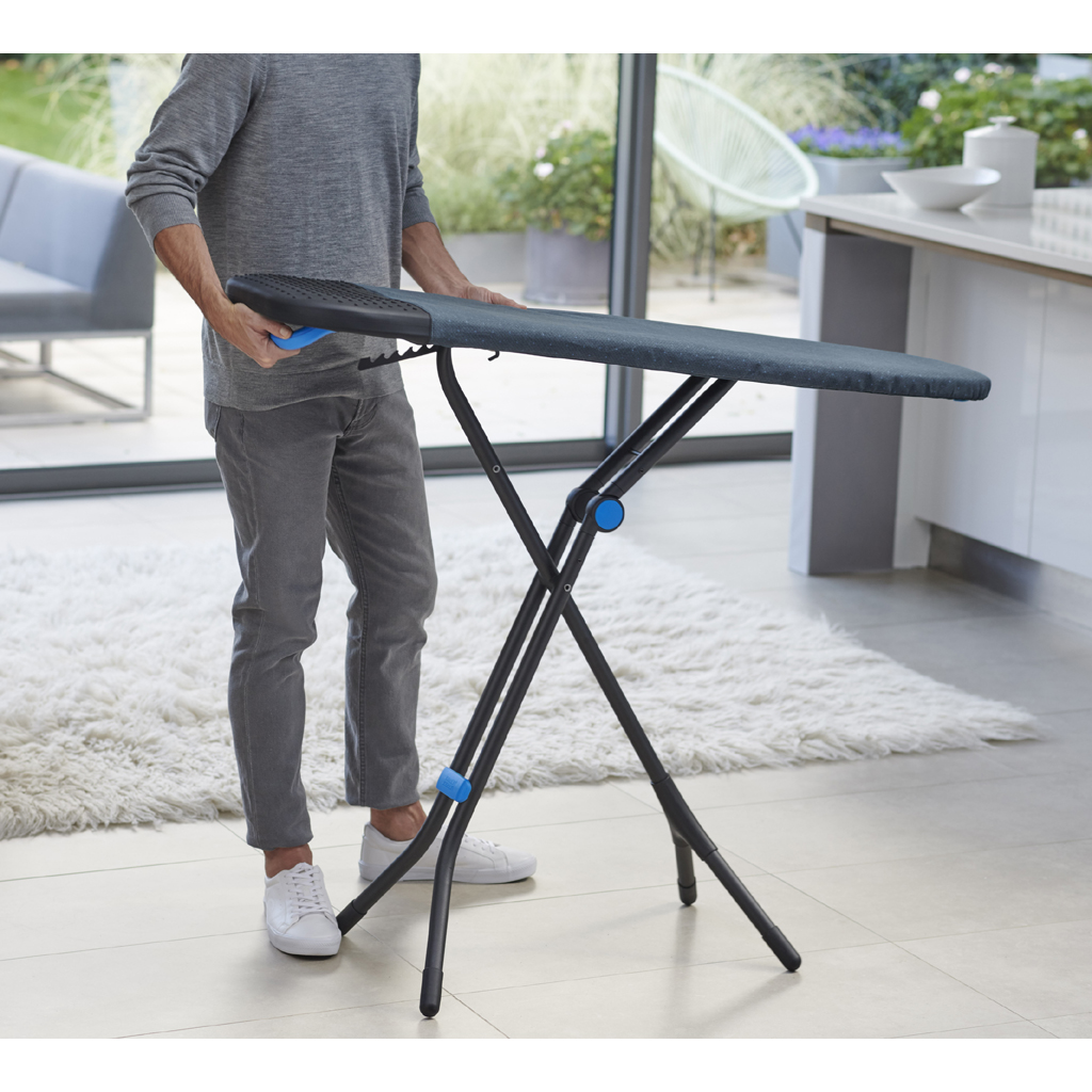 Un homme tient une table à repasser. Elle est noire et bleue. Il porte des vêtements gris et des chaussures blanches, dans une maison moderne.