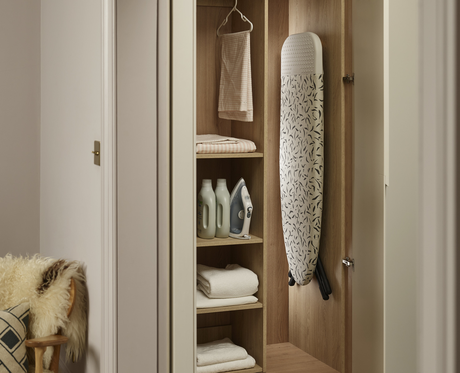 Ein Wäsche-Schrank mit Bügelbrett, Handtüchern und Reinigungsmitteln. Ein Stuhl mit Fell ist im Vordergrund.