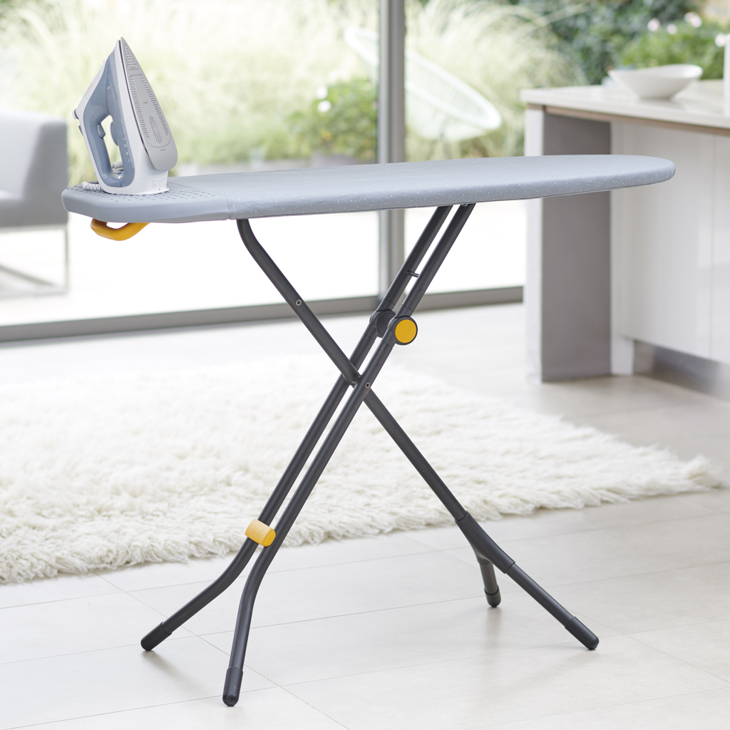 Une table à repasser avec un dessus gris et un fer à repasser argenté. La planche est sur un tapis blanc.