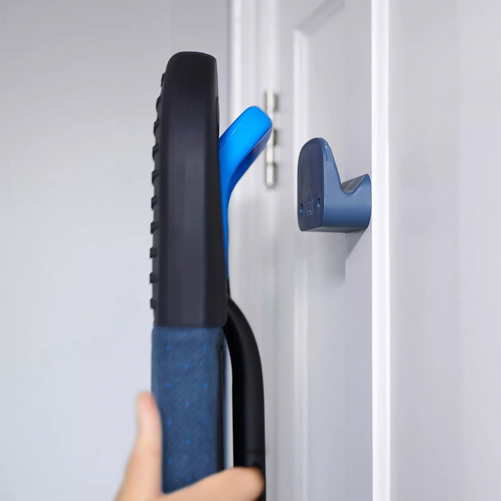 Un objet bleu et noir est suspendu à une porte blanche. Une main s'approche. Un crochet bleu est sur la porte.