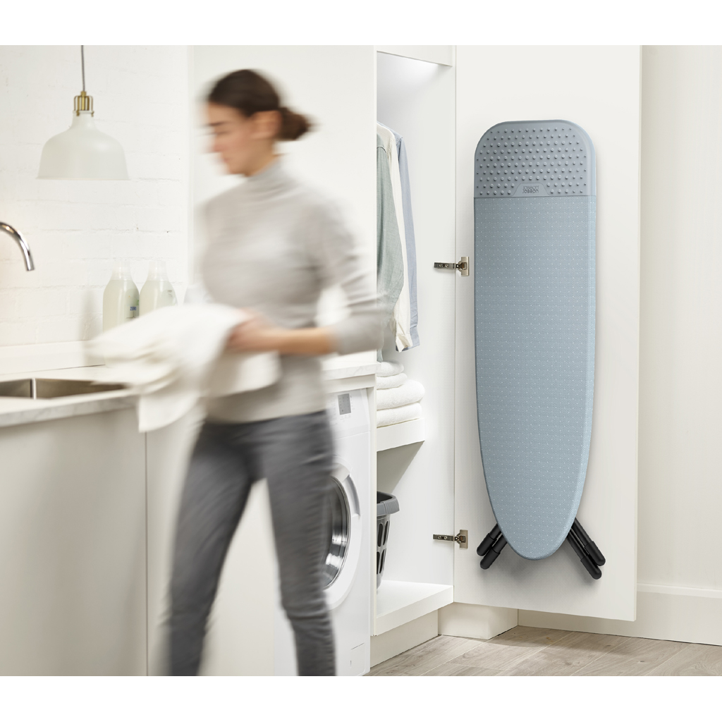 Une femme marche dans une buanderie, tenant des serviettes blanches. Une planche à repasser est rangée dans un placard. Machine à laver visible.