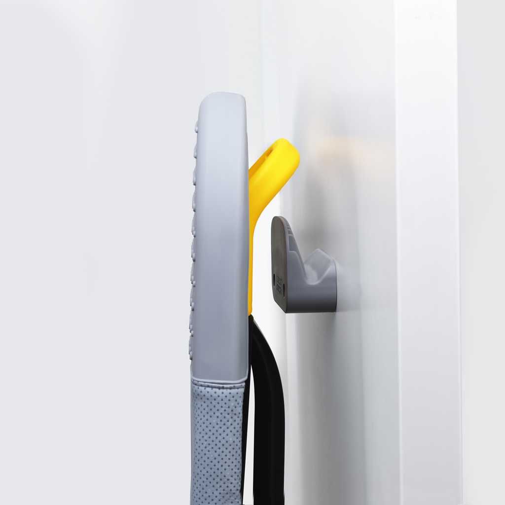 Une vadrouille grise et jaune est suspendue à un mur blanc. Un crochet gris maintient la vadrouille en place, et une poignée jaune est visible.
