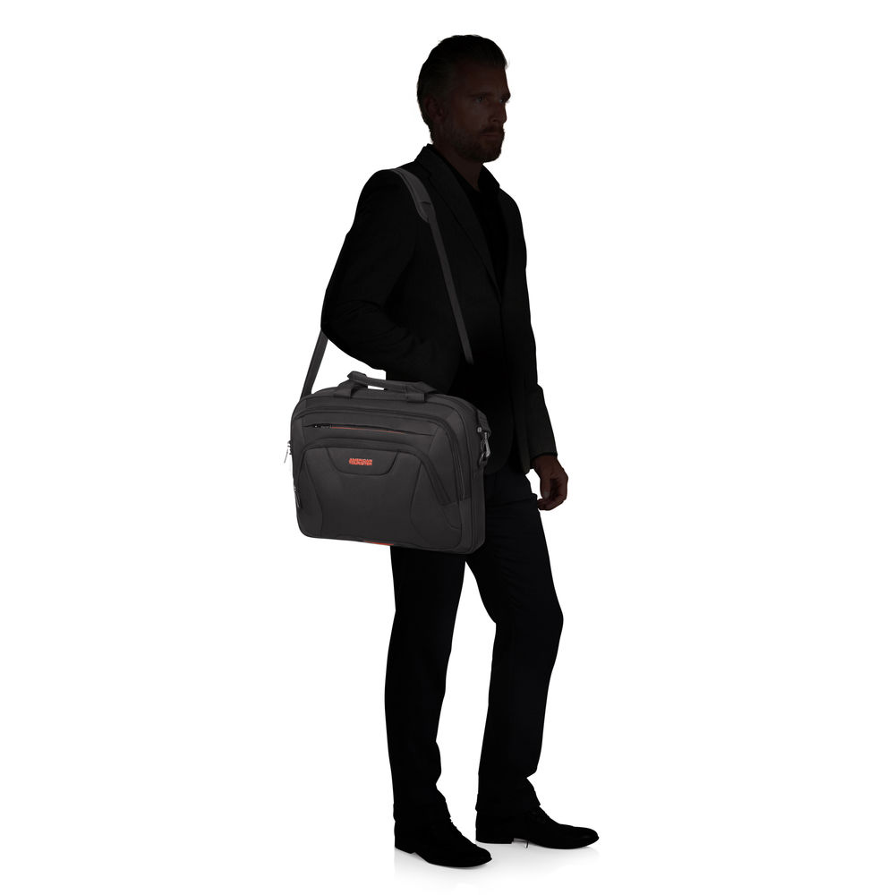 Un uomo in giacca e cravatta porta una borsa nera sulla spalla. La borsa ha un logo arancione. Lo sfondo è bianco.