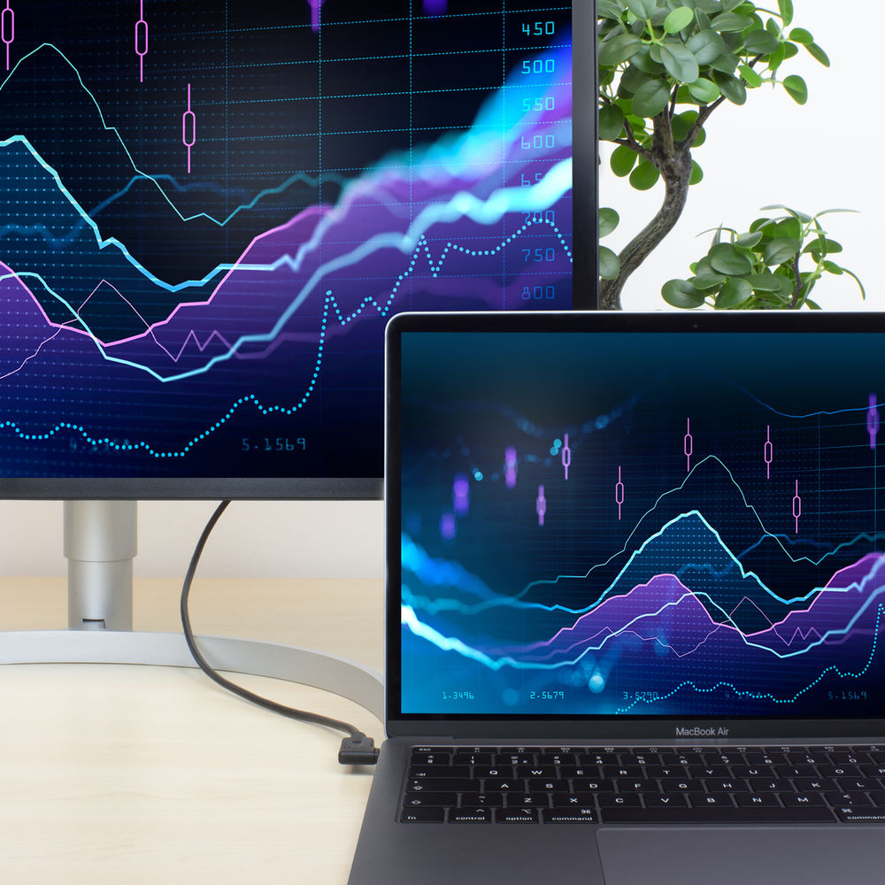 Een laptop en monitor tonen aandelenkoersen. Een kleine plant rechts. Een kabel verbindt de laptop met de monitor.