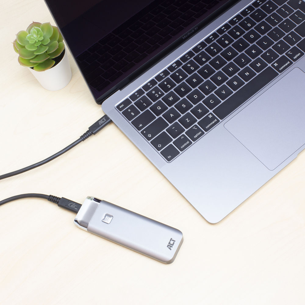 Een laptop is via een zwarte kabel verbonden met een zilveren externe harde schijf. Een plant in een witte pot links.