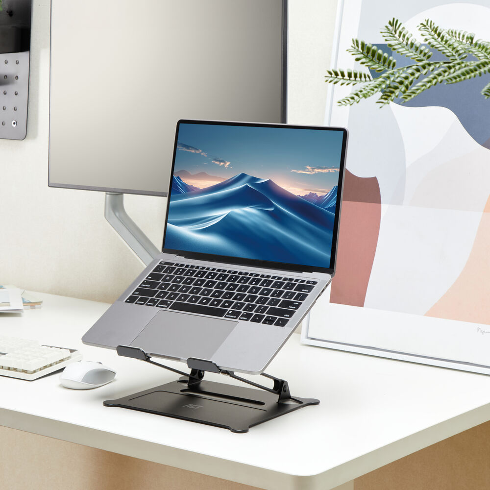 Ein Laptop auf einem Ständer zeigt eine Landschaft. Im Hintergrund ein Monitor und ein Kunstwerk. Die Szene spielt auf einem weißen Schreibtisch.