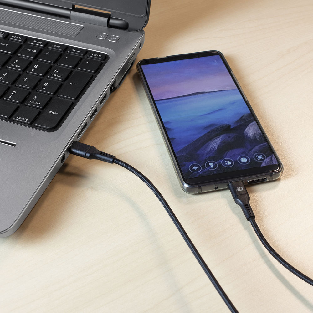 Un smartphone se recharge depuis un ordinateur portable avec un câble noir. Le téléphone affiche une photo de paysage. Les deux sont sur du bois.