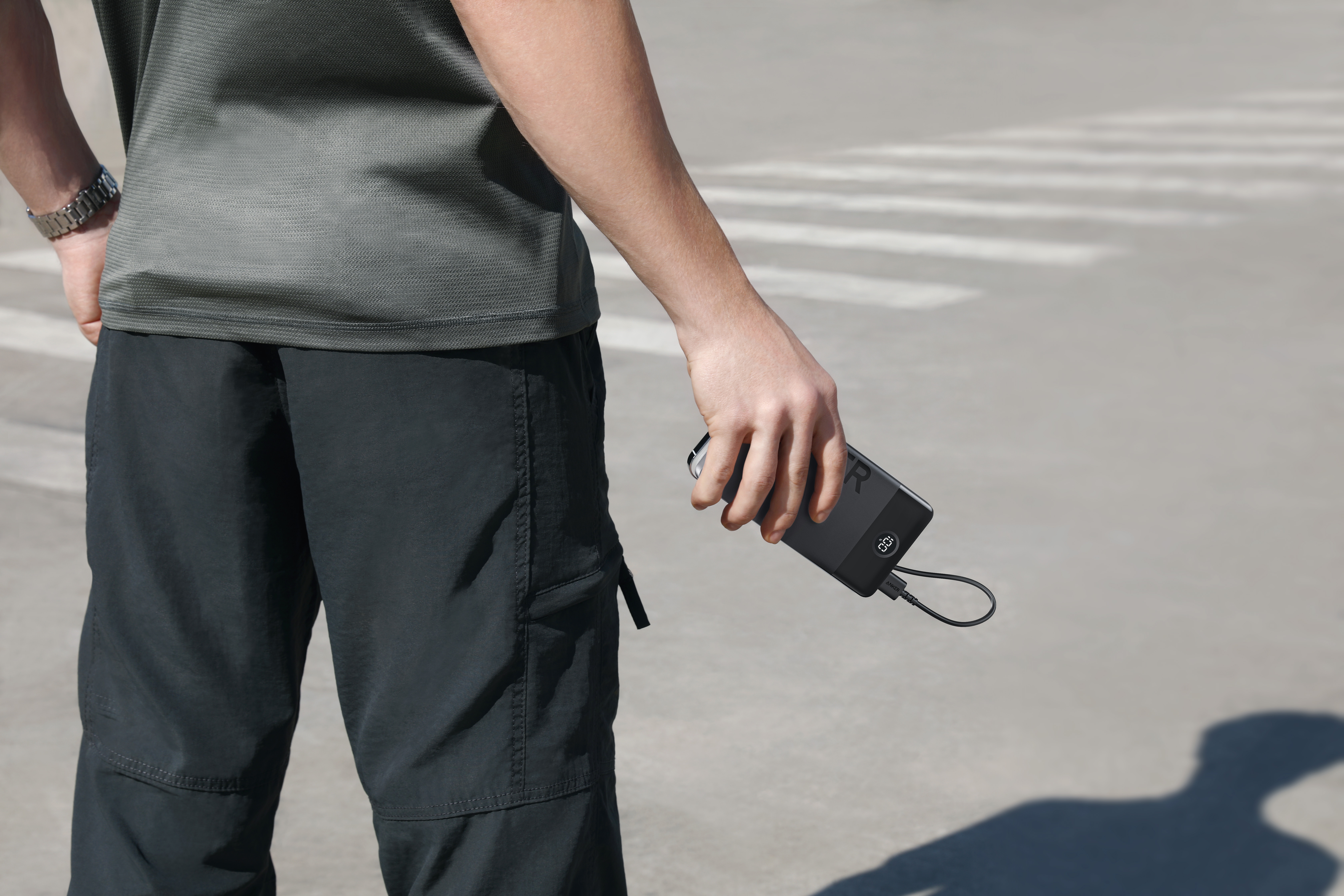 Persona sosteniendo un banco de energía negro con un cable de carga. Viste camisa gris y pantalones negros en un paso de cebra.