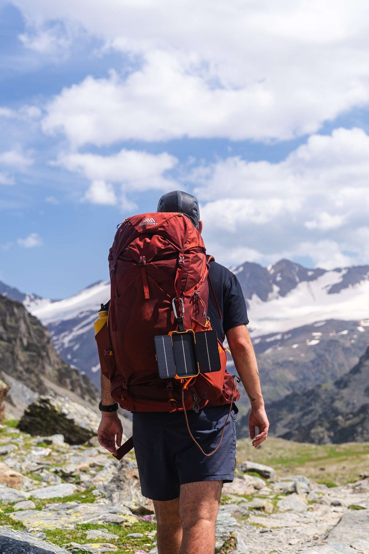 Ein Wanderer mit rotem Rucksack geht auf Berge zu. Solarpanel ist angebracht. Der Himmel ist blau mit weißen Wolken.