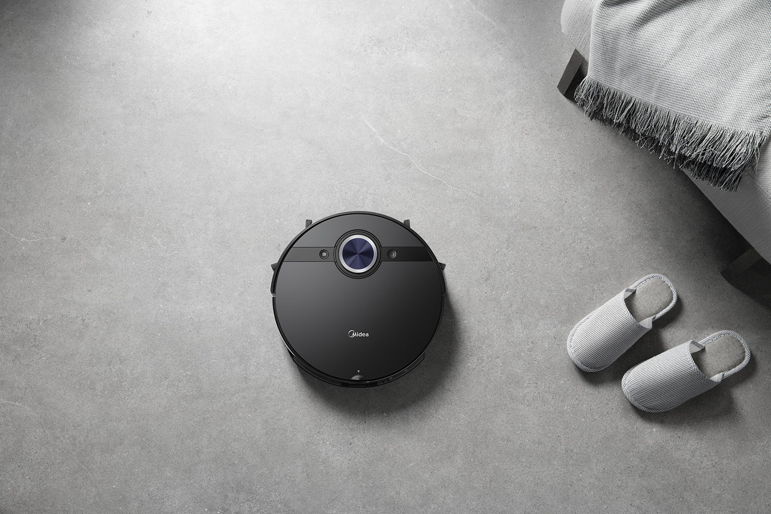 Un aspirateur robot noir, des pantoufles grises et une partie d'un canapé avec une couverture sur un sol gris.