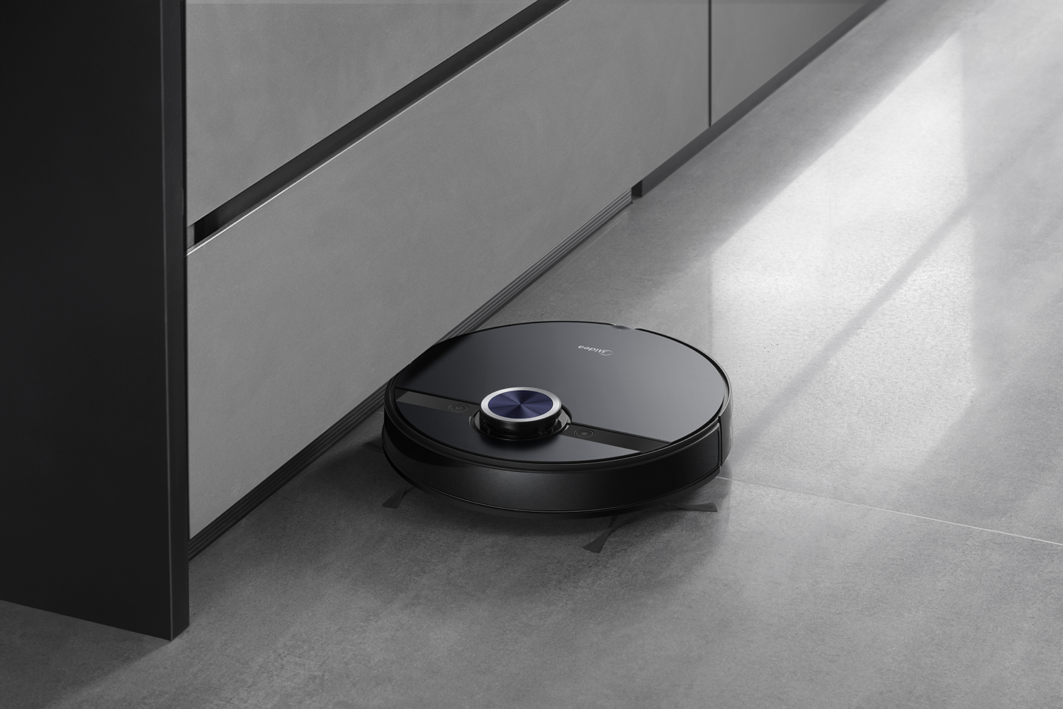 Un aspirateur robot noir sur un sol carrelé gris, près d'un meuble gris. L'aspirateur a un bouton bleu et le logo Midea.