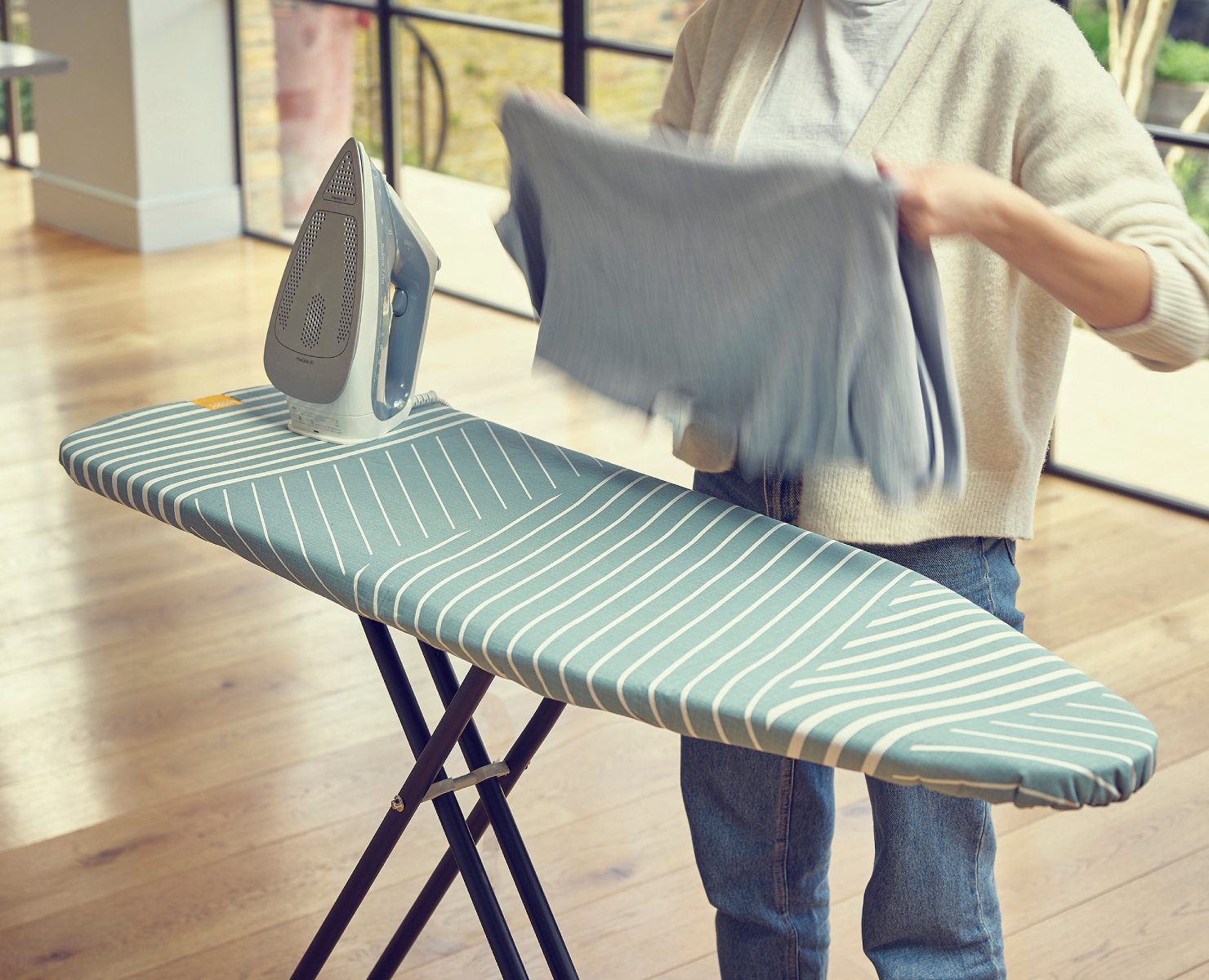 Una persona plancha ropa en una tabla de planchar. Una plancha está sobre la tabla. La tabla tiene un patrón de rayas azules y blancas.