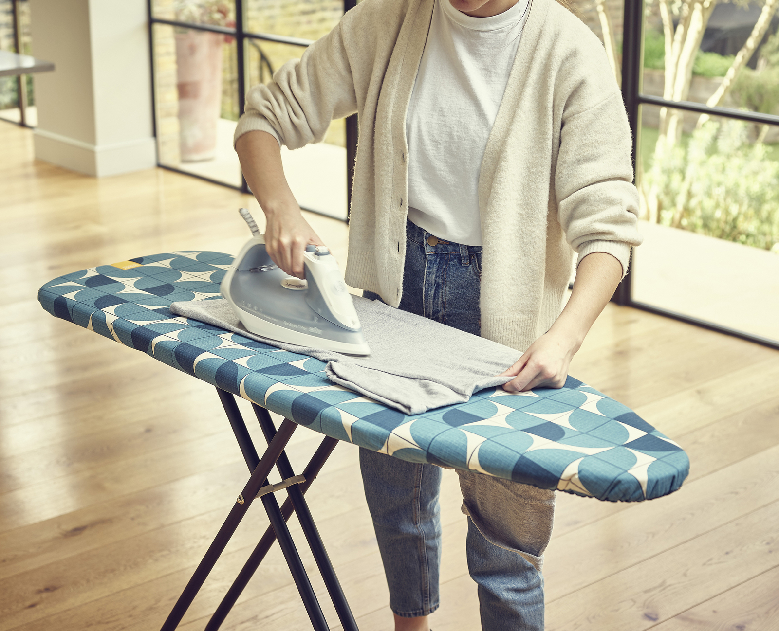 Una persona plancha ropa gris en una tabla de planchar estampada. Viste jeans y un cárdigan. Ventanas y suelo de madera al fondo.
